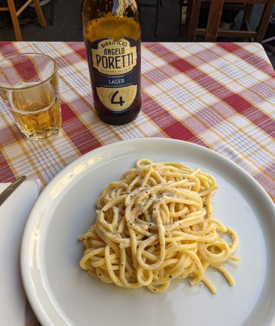 Cacio e pepe pasta served on a white plate with a glass and bottle of Angelo Poretti lager on a checkered tablecloth at Trattoria Luzzi in Rome.