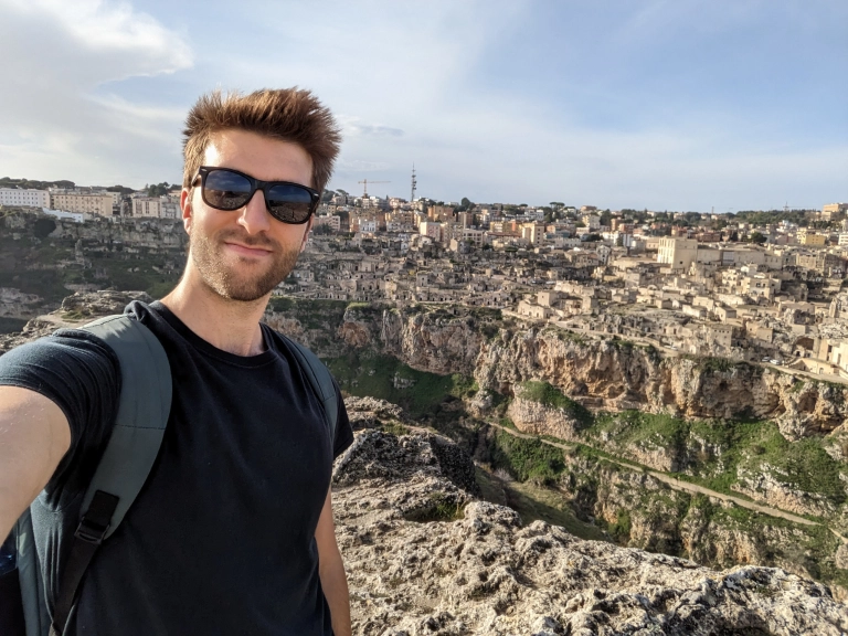 A man poses with the ancient stone city of Matera behind him, wearing a black t-shirt, sunglasses, and a backpack.