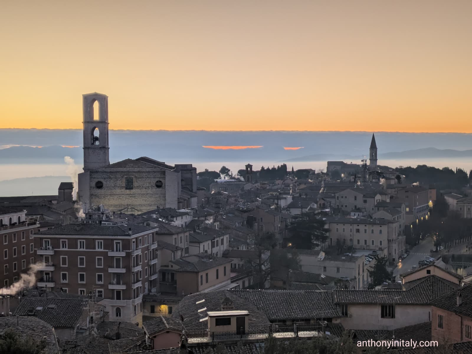 Sunrise over the rooftops of Perugia, Umbria, with the Church of San Domenico and other historic towers silhouetted against a soft orange sky.