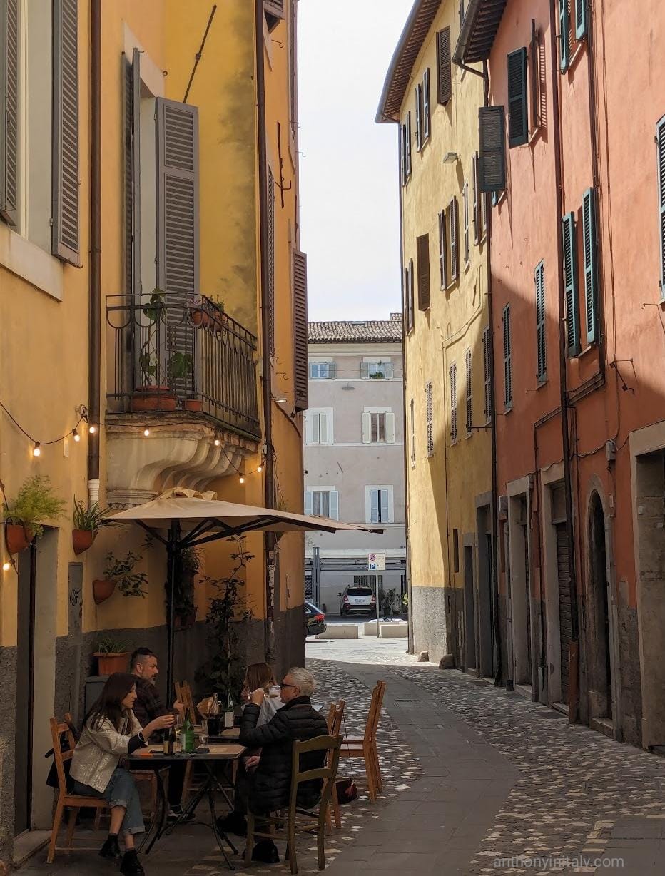 Narrow cobblestone street in an Italian town with colorful buildings, open shutters, and a small outdoor café where people sit under an umbrella enjoying drinks and conversation.