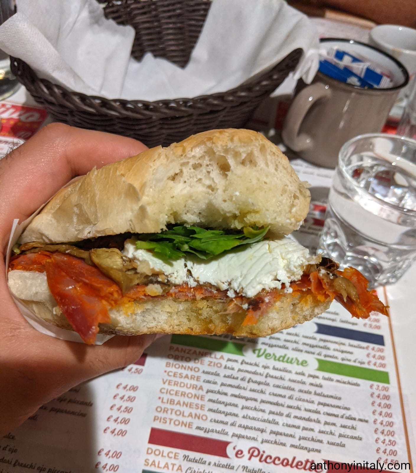 Half-eaten sandwich from Dar Ciriola in Rome, filled with salame piccante (or ventricina), grilled eggplant, leafy greens, and a generous spread of creamy cheese, held over a printed menu.