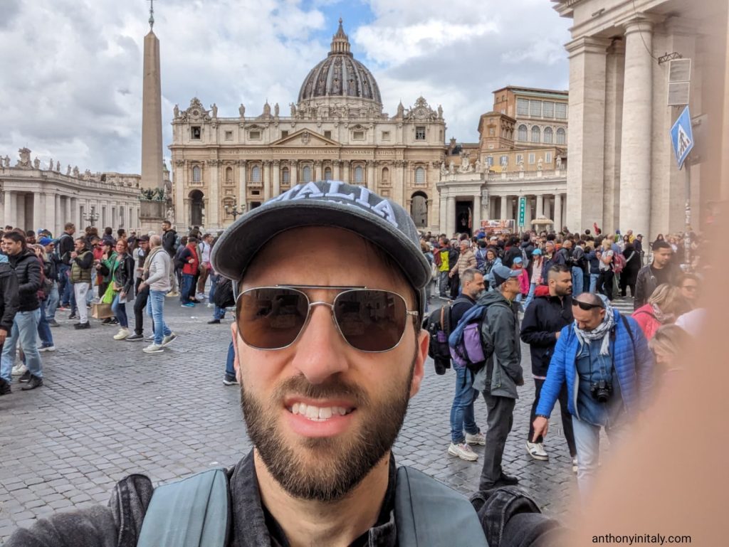 Man wearing sunglasses and a baseball cap that says ‘ITALIA’ taking a selfie in St. Peter’s Square, Vatican City, with a crowd of tourists and St. Peter’s Basilica visible in the background under a partly cloudy sky