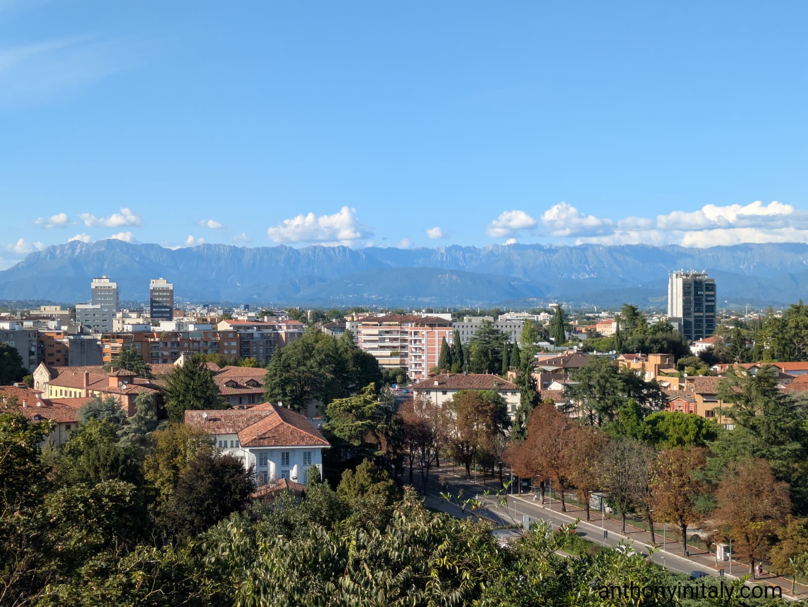Panoramic view of Udine with red-tiled roofs, modern buildings, and the Julian Alps in the background on a clear day