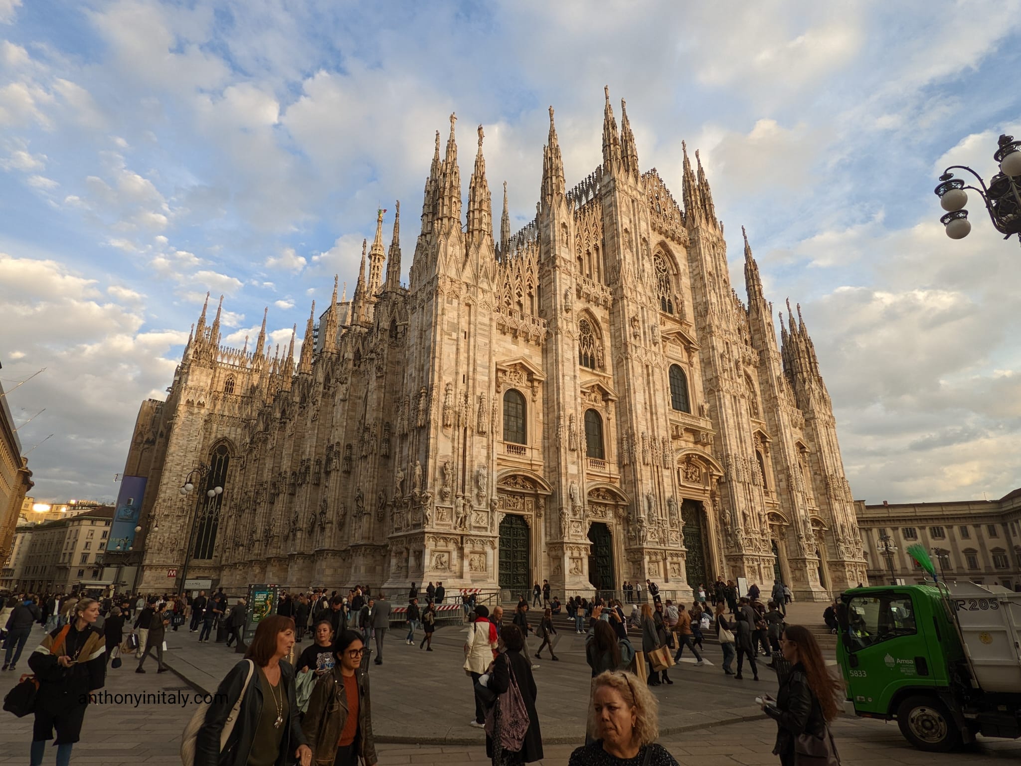 The facade of Milan's Duomo cathedral at golden hour, with crowds of people crossing the piazza in front of it.
