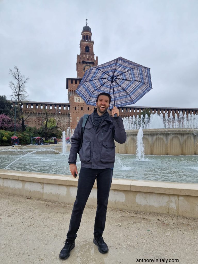 Man smiling and holding a plaid umbrella in front of the Castello Sforzesco fountain in Milan on a cloudy day.