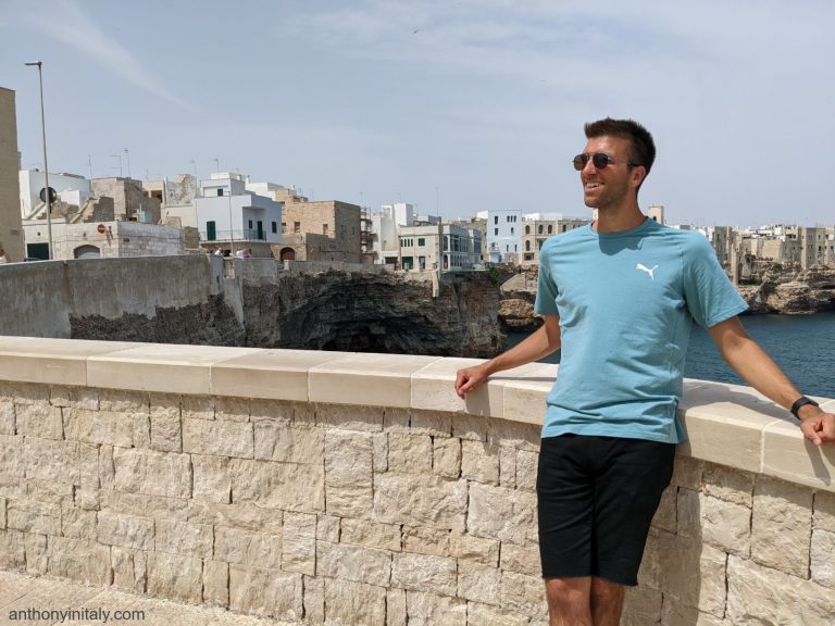 Anthony from Anthony in Italy standing on a stone bridge overlooking the cliffs and turquoise water of Polignano a Mare in Puglia, with the town's white and stone buildings visible in the background.