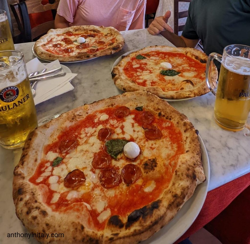 Three Neapolitan pizzas on a marble table at Pizzeria I Decumani in Naples: two Diavola pizzas with spicy salami and buffalo mozzarella, and one classic Margherita, served with tall beers.