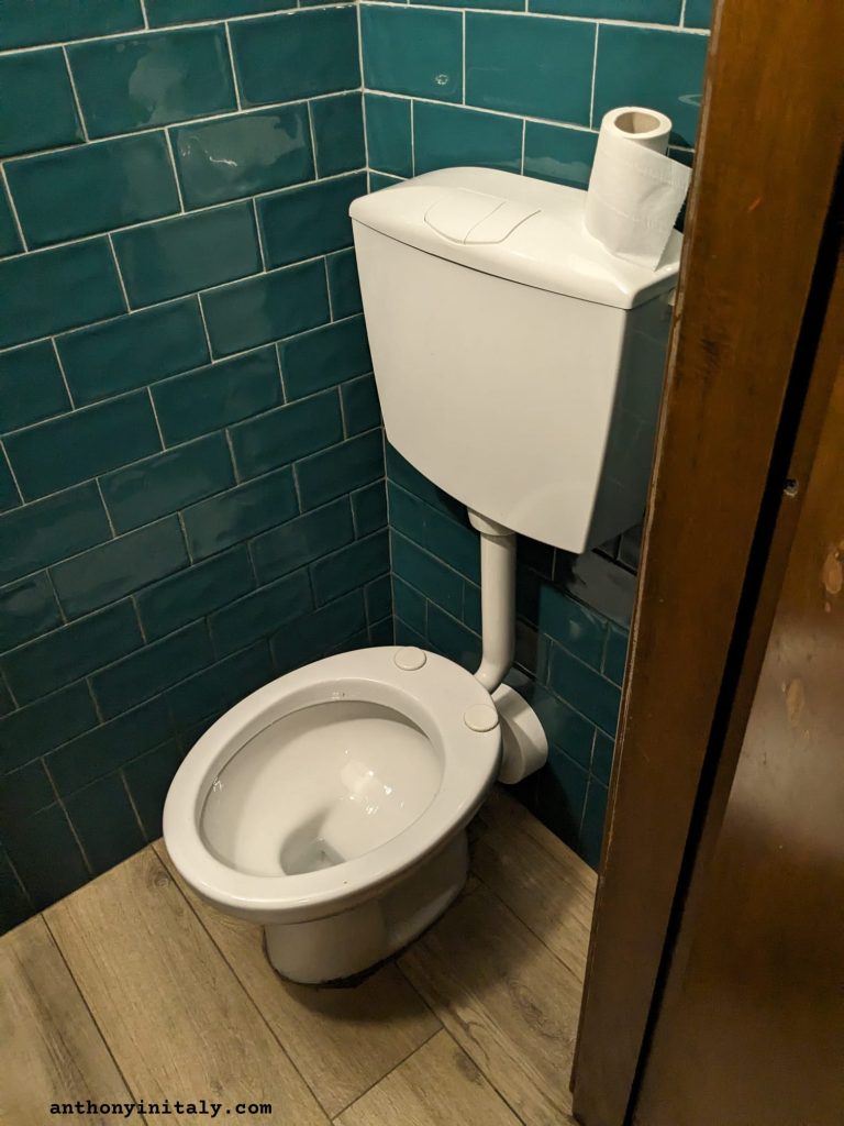 A public toilet without a seat in an Italian bar, set against teal tiles, photographed by Anthony in Italy
