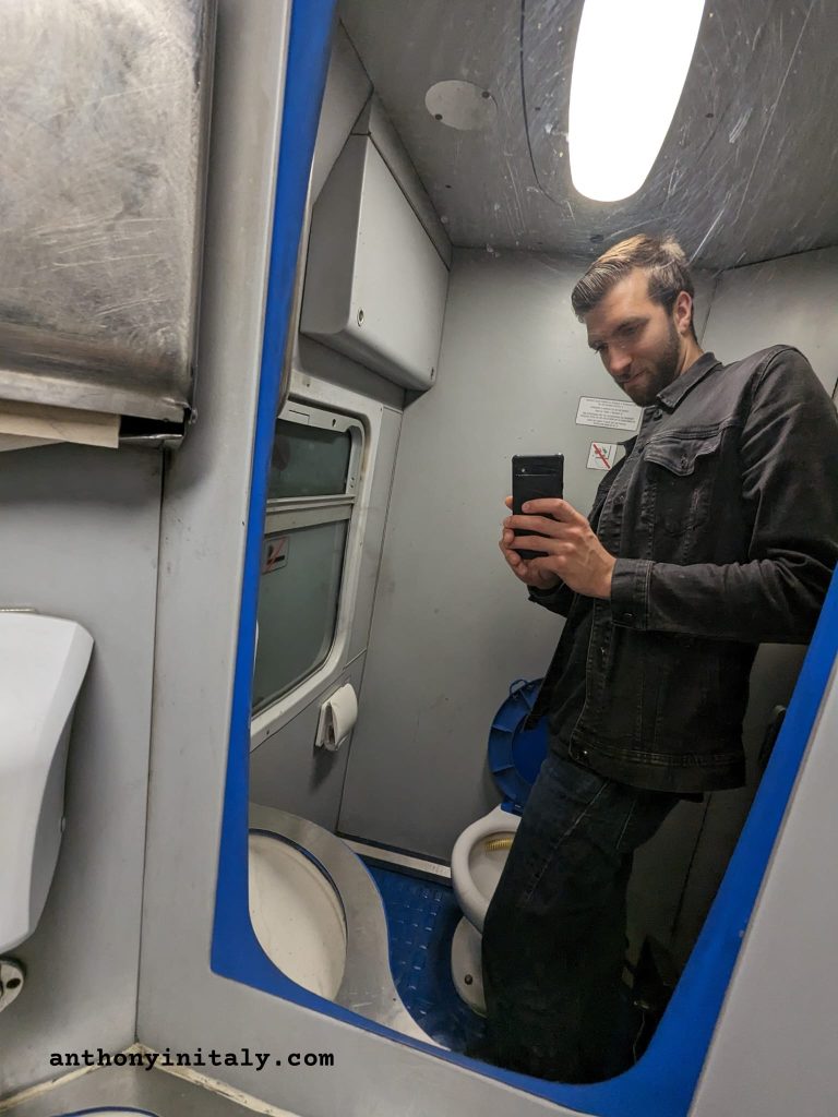 Anthony in Italy taking a mirror selfie inside an Italian regional train bathroom, showing the blue and grey interior with toilet visible in the reflection