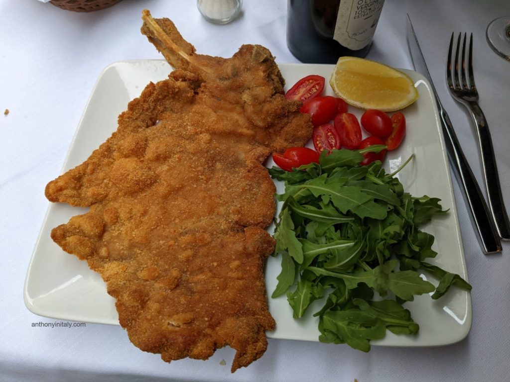 A large bone-in cotoletta alla Milanese — breaded and pan-fried veal cutlet — served with arugula and cherry tomatoes
