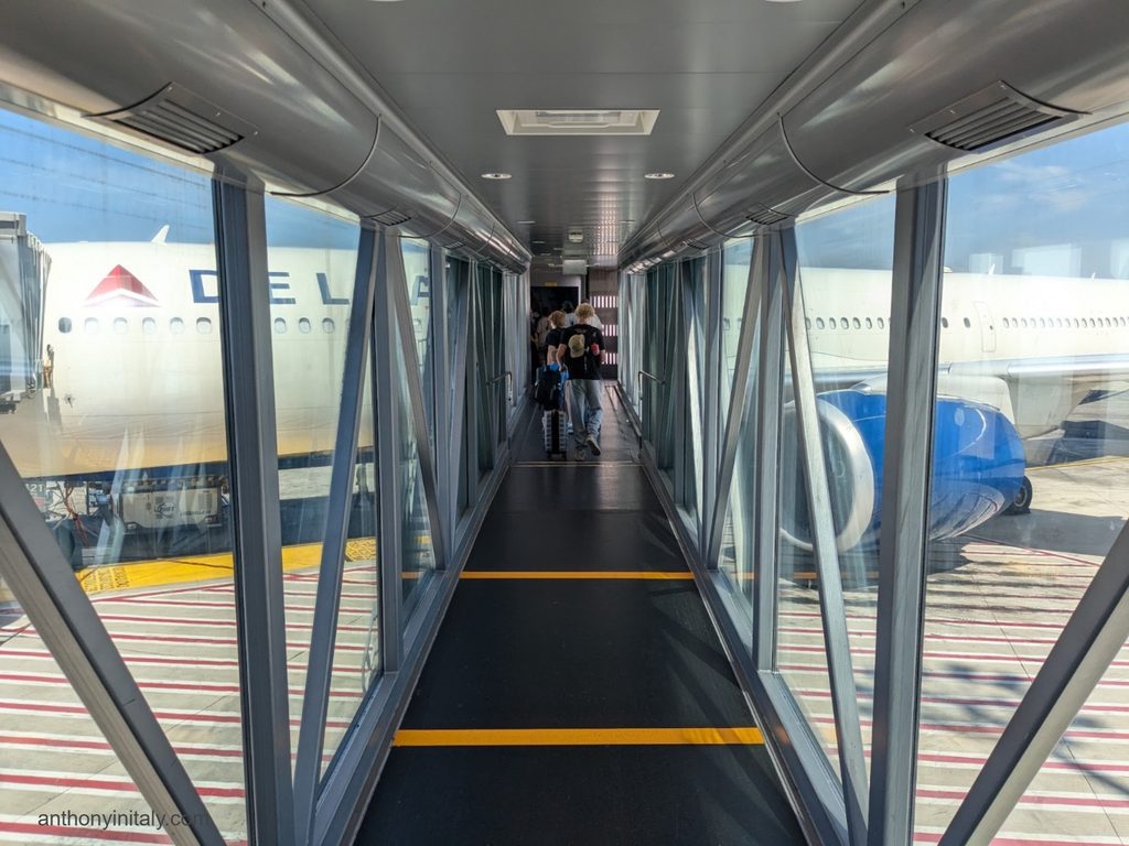 View down an airport jet bridge toward a waiting Delta airplane, with passengers walking toward the aircraft door.