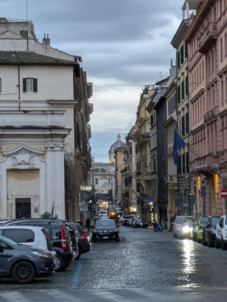 An overcast morning view down a narrow Roman street near Piazza di San Bernardo, with parked cars, illuminated shop and hotel signs, and a church dome visible in the distance