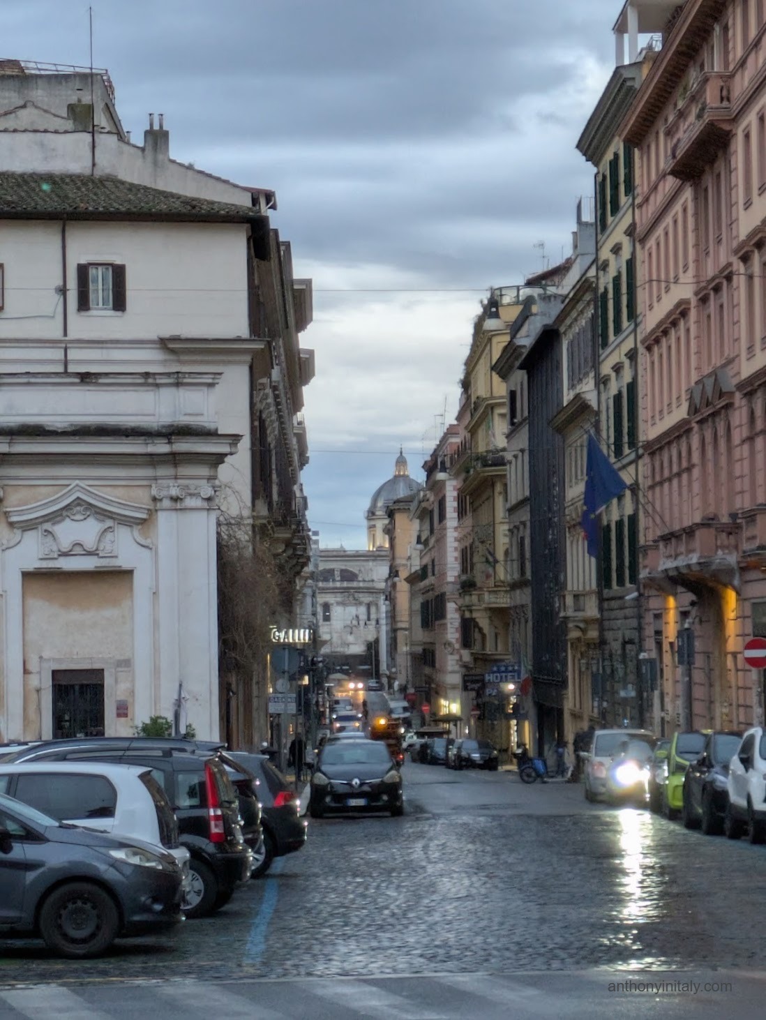 An overcast morning view down a narrow Roman street near Piazza di San Bernardo, with parked cars, illuminated shop and hotel signs, and a church dome visible in the distance