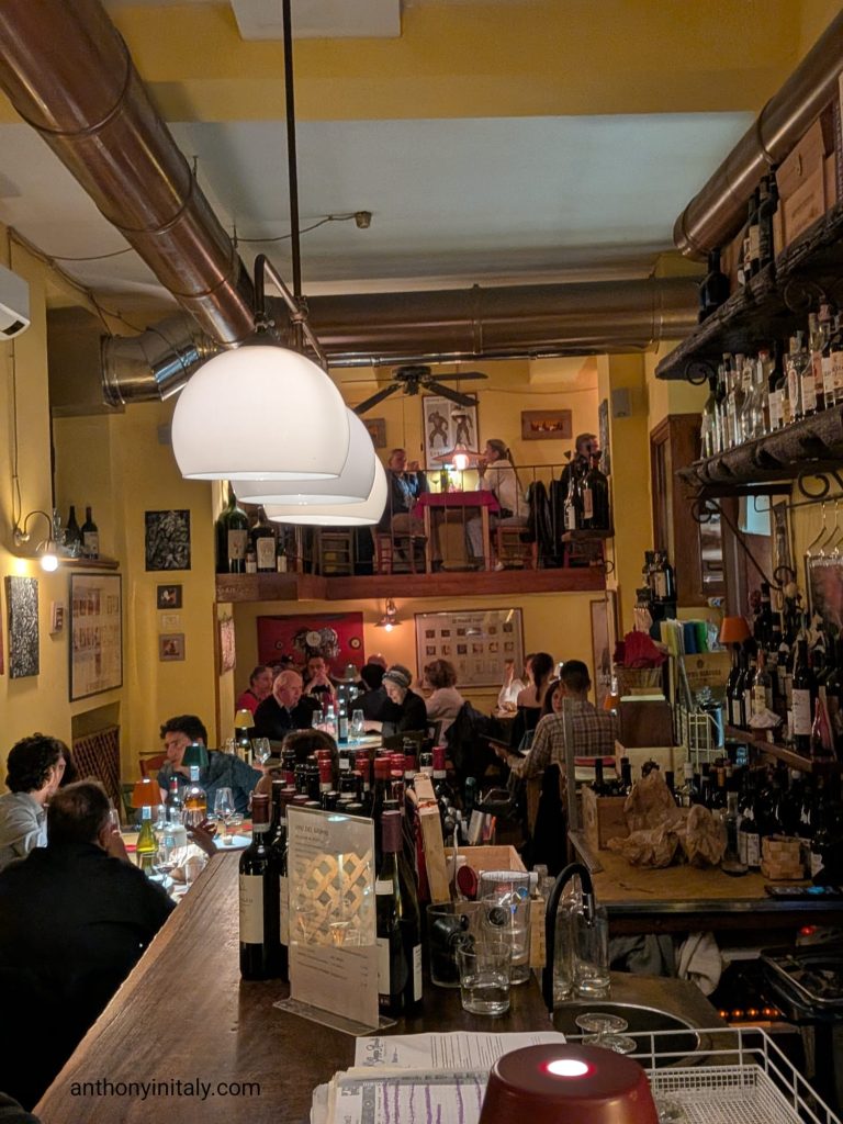 Warm, yellow-walled dining room at Civico 25 in Perugia with a wine-lined bar, packed tables, and a small loft level overlooking the room